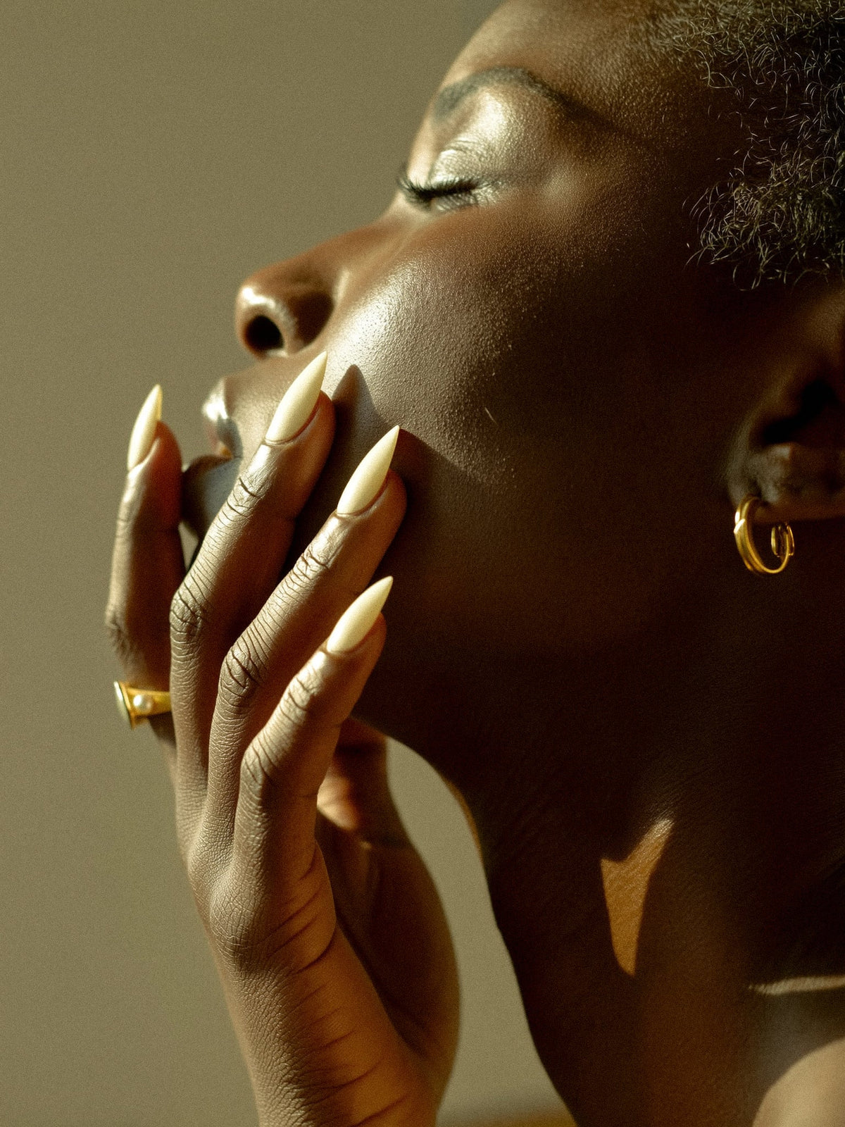 Profile of a woman with eyes closed and her hand at her lips, with long nails, a gold ring, and a gold hoop earring in warm light