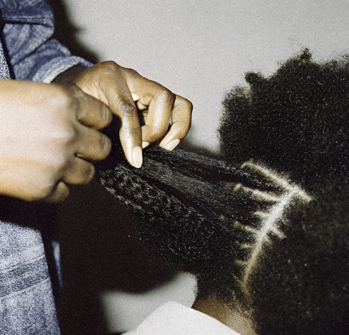 A close-up lifestyle shot of a person braiding another person's hair. The braider's hands are actively weaving a section of dark hair into a braid, while the recipient has visible light-colored hair streaks or highlights running through the dark hair.
