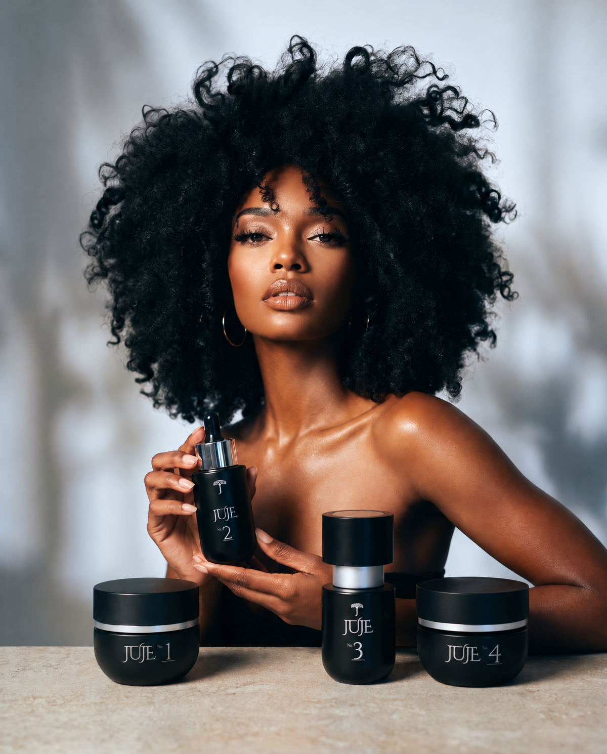Model with a voluminous afro holding the Juje No. 2 Scalp Serum, with No. 1, No. 3 and No. 4 on the table in front of her