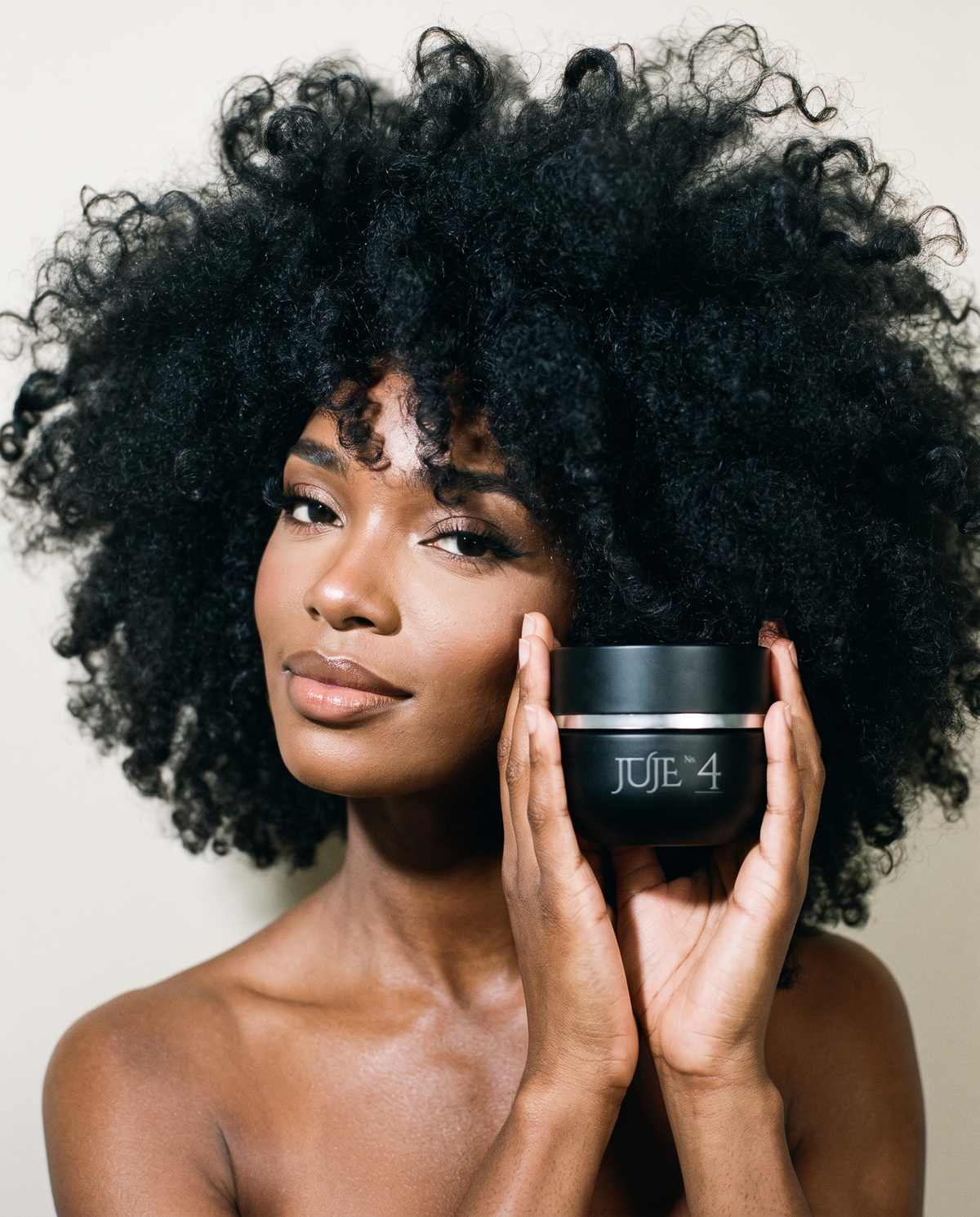 Model with voluminous curly hair holding the Juje No. 4 Hair Masque jar to her cheek and looking at the camera